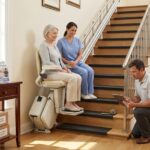Older adult testing a stairlift at home with an installer checking measurements and a caregiver watching